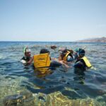 Swimmers in yellow vests, masks and snorkels in the clear water of the Blue Hole, with the Sinai mountains behind